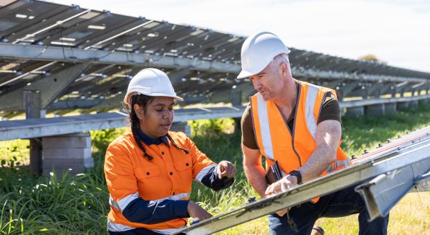 A colour photo of two workers, a man and a woman, discussing solar panels at a solar plant. They are wearing white hats and orange high-visibility shirts.