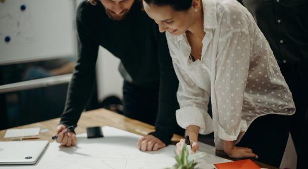 A colour photo of two workers, a man and a woman, in an office looking at plans on an desk in a modern office.