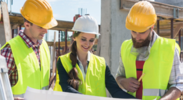 A colour photo of two men and a woman in bright yellow construction attire looking at a building plan in a construction zone.