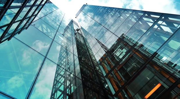 A colour photo from the perspective of looking up at a glass office building and the blue sky with clouds. The building is modern and corporate.