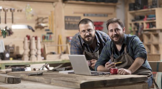 A colour photo of two men looking at the viewer and smiling. They are working on a laptop in a workshop.