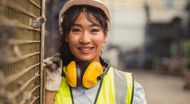 A colour photo of a woman wearing a white hard hat, work gloves, a yellow hi-vis vest and headphones are around her neck. 