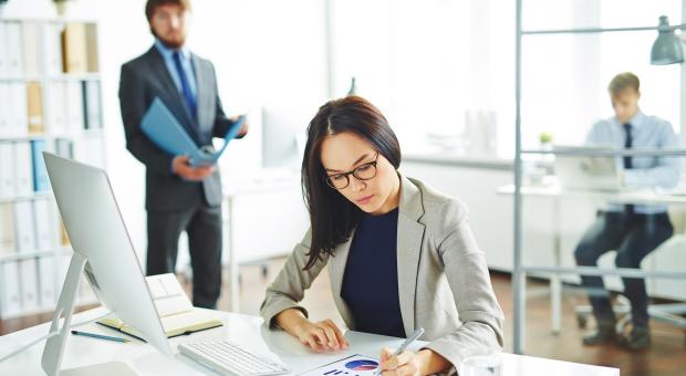 A photo of a woman sitting at a desk with a computer and keyboard, looking at a document on the desk. A man is in the background walking towards the woman. 