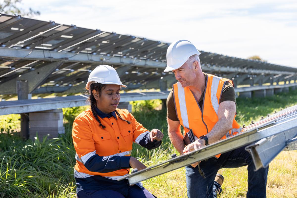 A colour photo of two workers, a man and a woman, discussing solar panels at a solar plant. They are wearing white hats and orange high-visibility shirts.