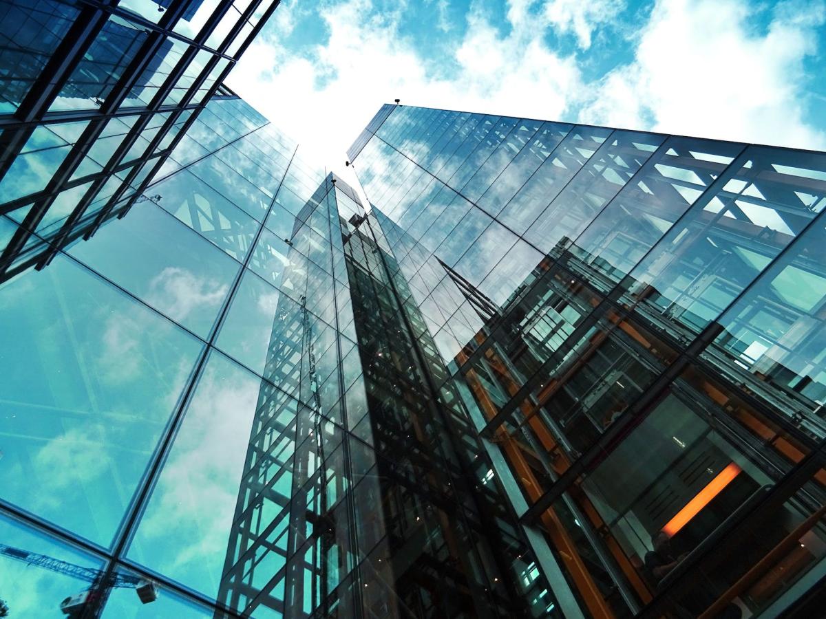 A colour photo from the perspective of looking up at a glass office building and the blue sky with clouds. The building is modern and corporate.