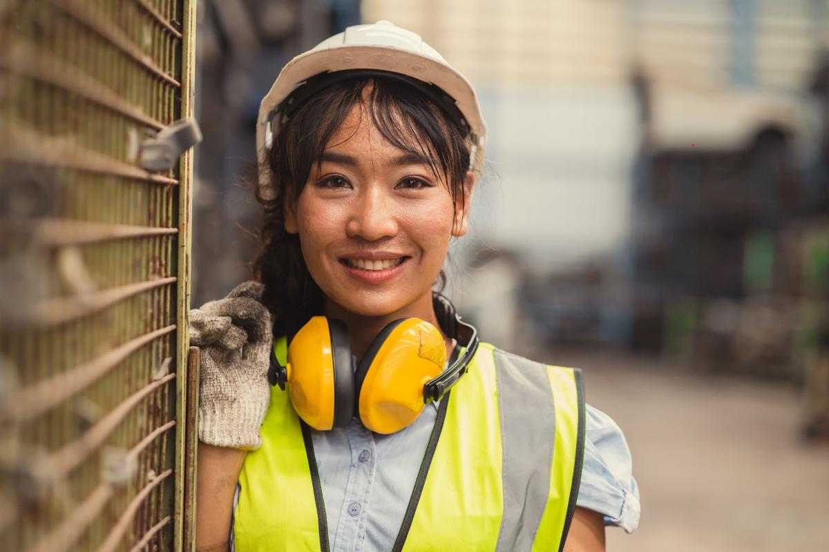 A colour photo of a woman wearing a white hard hat, work gloves, a yellow hi-vis vest and headphones are around her neck. 