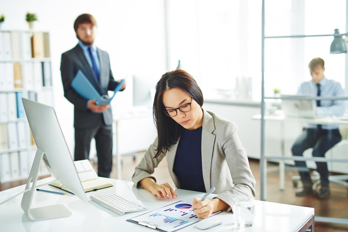 A photo of a woman sitting at a desk with a computer and keyboard, looking at a document on the desk. A man is in the background walking towards the woman. 